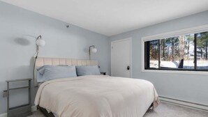 Um quarto com cama, mesas de cabeceira, janela com vista para árvores e uma porta.