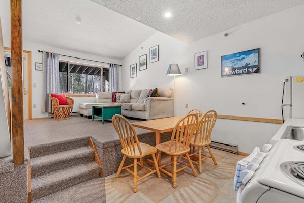 A cozy living area with a sofa, a dining table with chairs, a lamp, and a refrigerator.