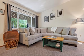 Ein Wohnzimmer mit einem Ecksofa, einem Holztisch, einem Holzstuhl und einer Stehlampe. An der Wand hängen drei gerahmte Bilder.
