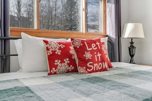 Een bed met een sprei, twee kussens met sneeuwvlindes, een lamp en een raam met uitzicht op bomen.