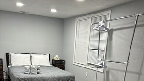A bedroom with a bed, a nightstand, a window with blinds, and a metal rack.