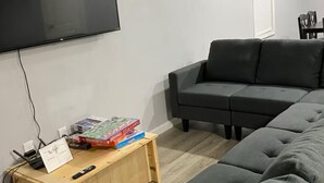 A modern living room with a grey sectional sofa, a wooden coffee table with books, a flat-screen TV mounted on the wall, and a window with blinds.