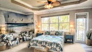 Ein Schlafzimmer mit einem großen Bett, zwei Nachttischen, einem Deckenventilator, einem Fenster mit Aussicht und einem Wandgemälde mit einer Strandansicht.