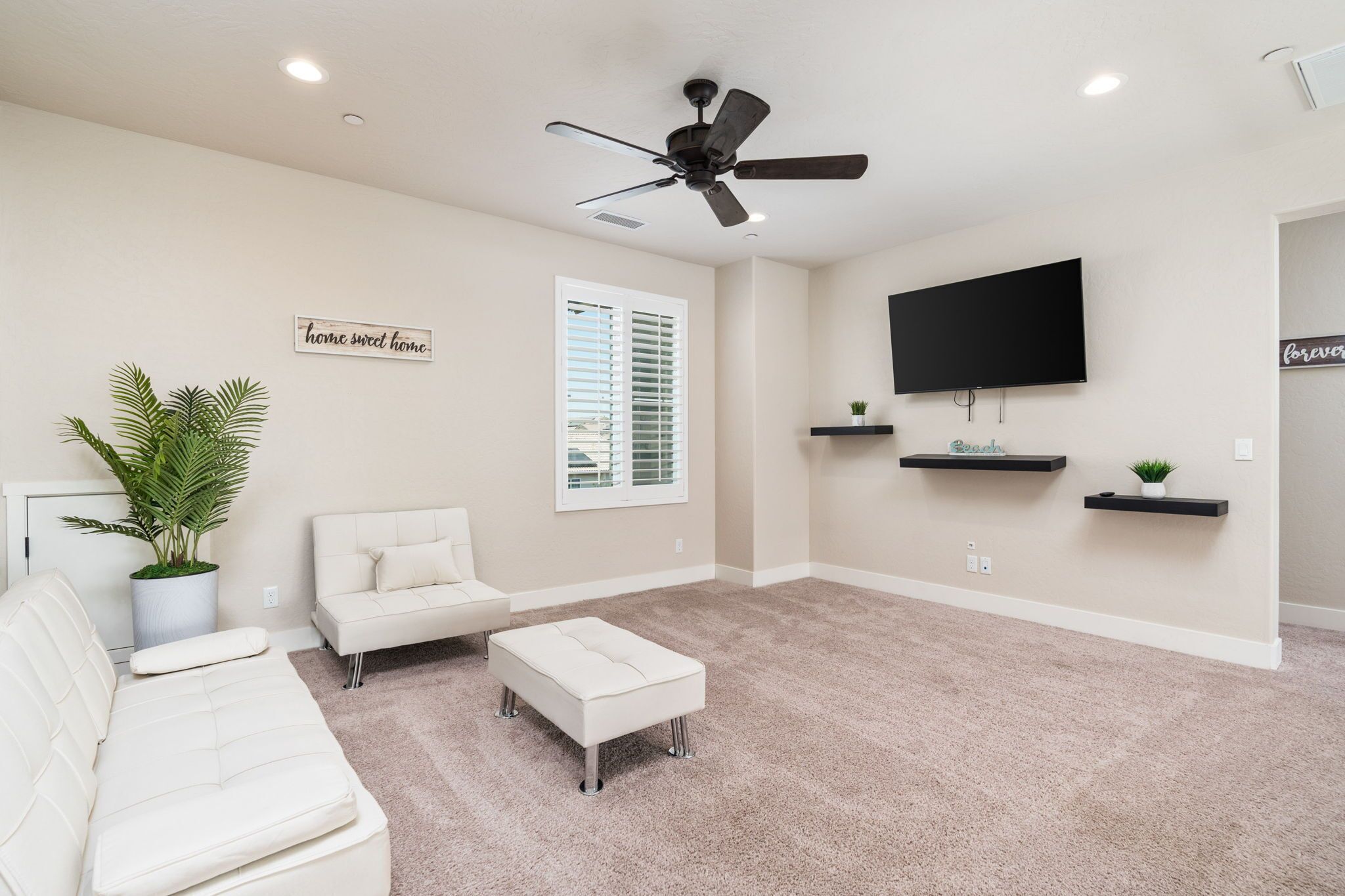 A living room with a beige carpet, white sofas, a ceiling fan, a flat-screen TV, and wall-mounted shelves with plants.