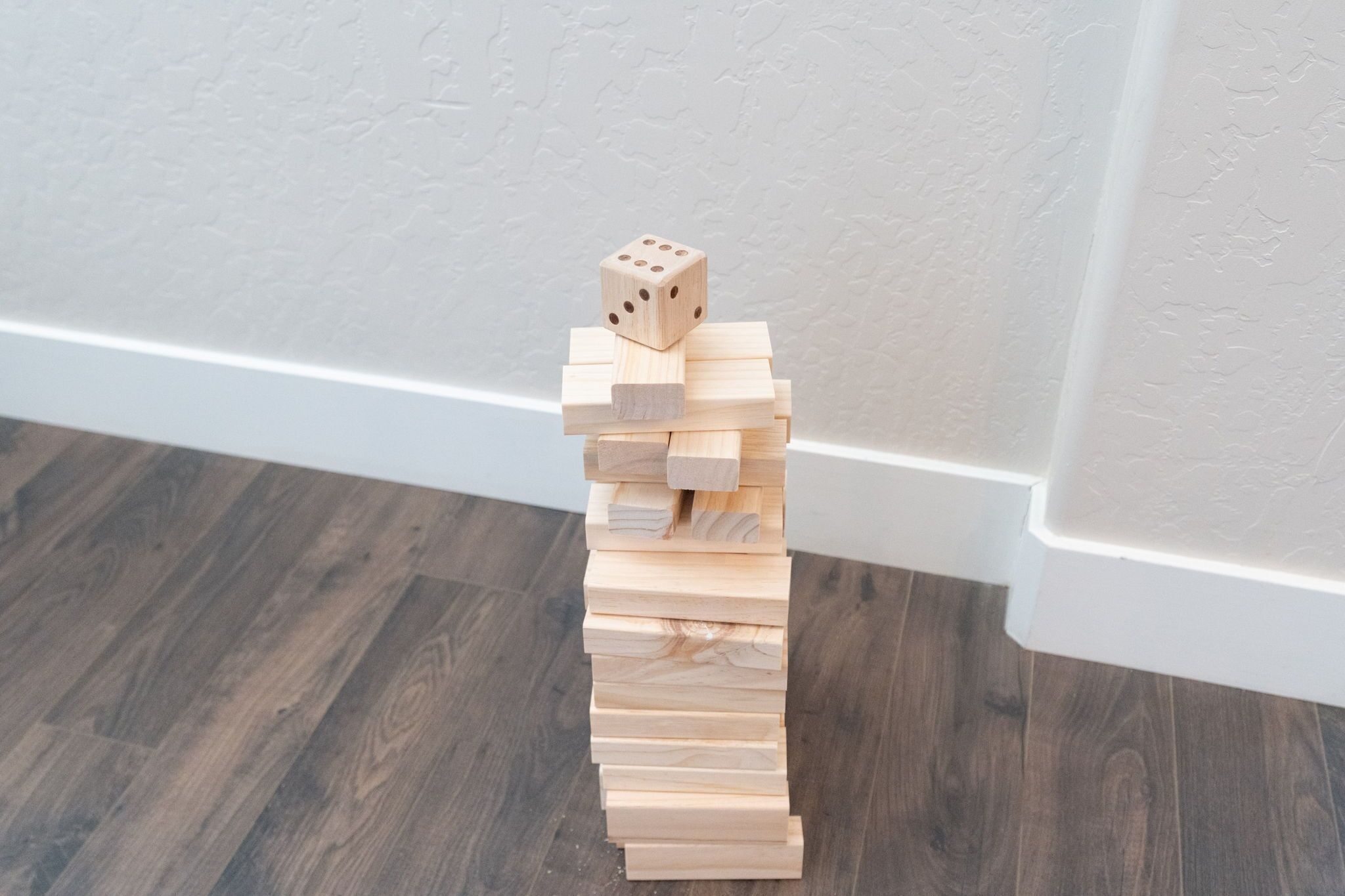 Ein Jenga-Turm aus Holz mit einem Würfel obenauf, auf einem Holzboden vor einer weißen Wand platziert.