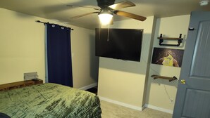 A bedroom with a bed, a ceiling fan, a TV mounted on the wall, a door with a towel rack, and a carpeted floor.
