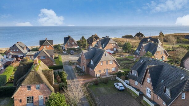 Eine Küstenwohngegend mit Häusern mit steilen Dächern und Backsteinfassade, umgeben von viel Grün und Blick auf das Meer.