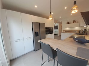 A modern kitchen with white cabinets, a stainless steel refrigerator, a wooden island with chairs, and pendant lighting.