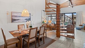 A dining area with a wooden table and chairs, a hanging light fixture, and a staircase leading to an upper level. - SUMMER SALE! Freshly Updated 2BR w Ocean Views - Kamaole Sands 8409 (Kihei)