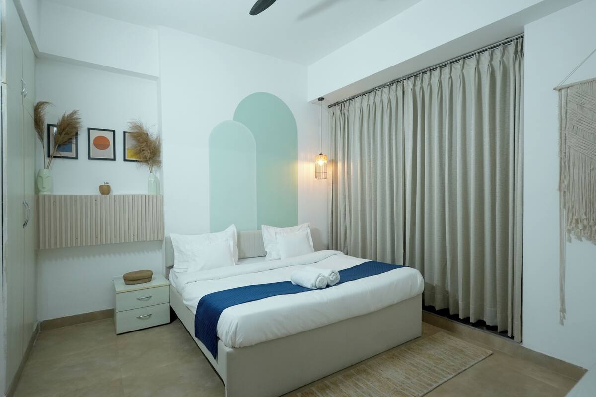 Una habitación de hotel moderna con una cama grande, una mesita de noche, un estante montado en la pared con adornos y una ventana con cortinas.