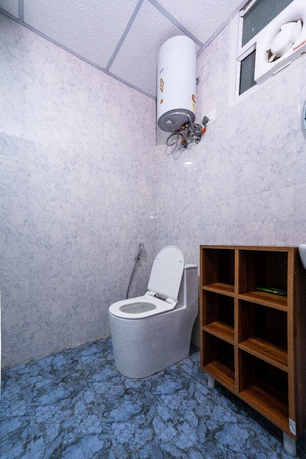 A bathroom with a toilet, a wooden shelf, and a water heater mounted on the wall. - Nimantrana (Noida)