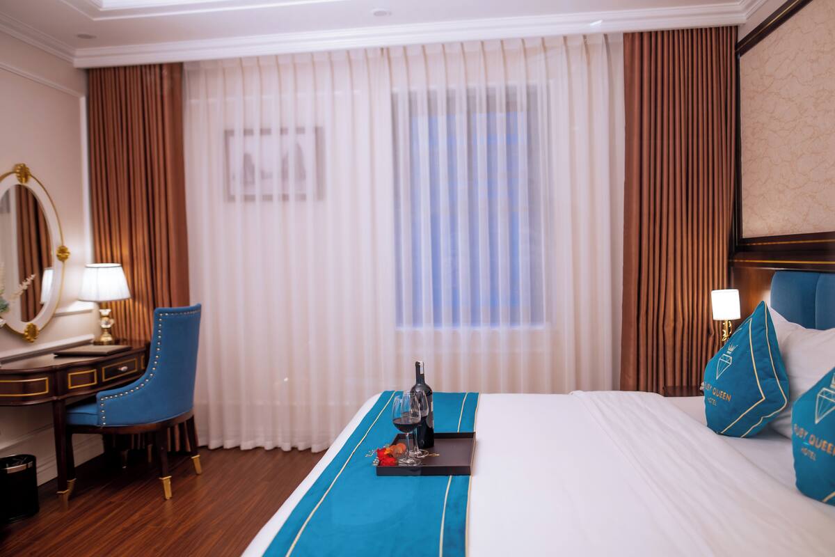 A hotel room with a bed, a desk, a chair, a window with curtains, and a tray with a bottle and glasses.
