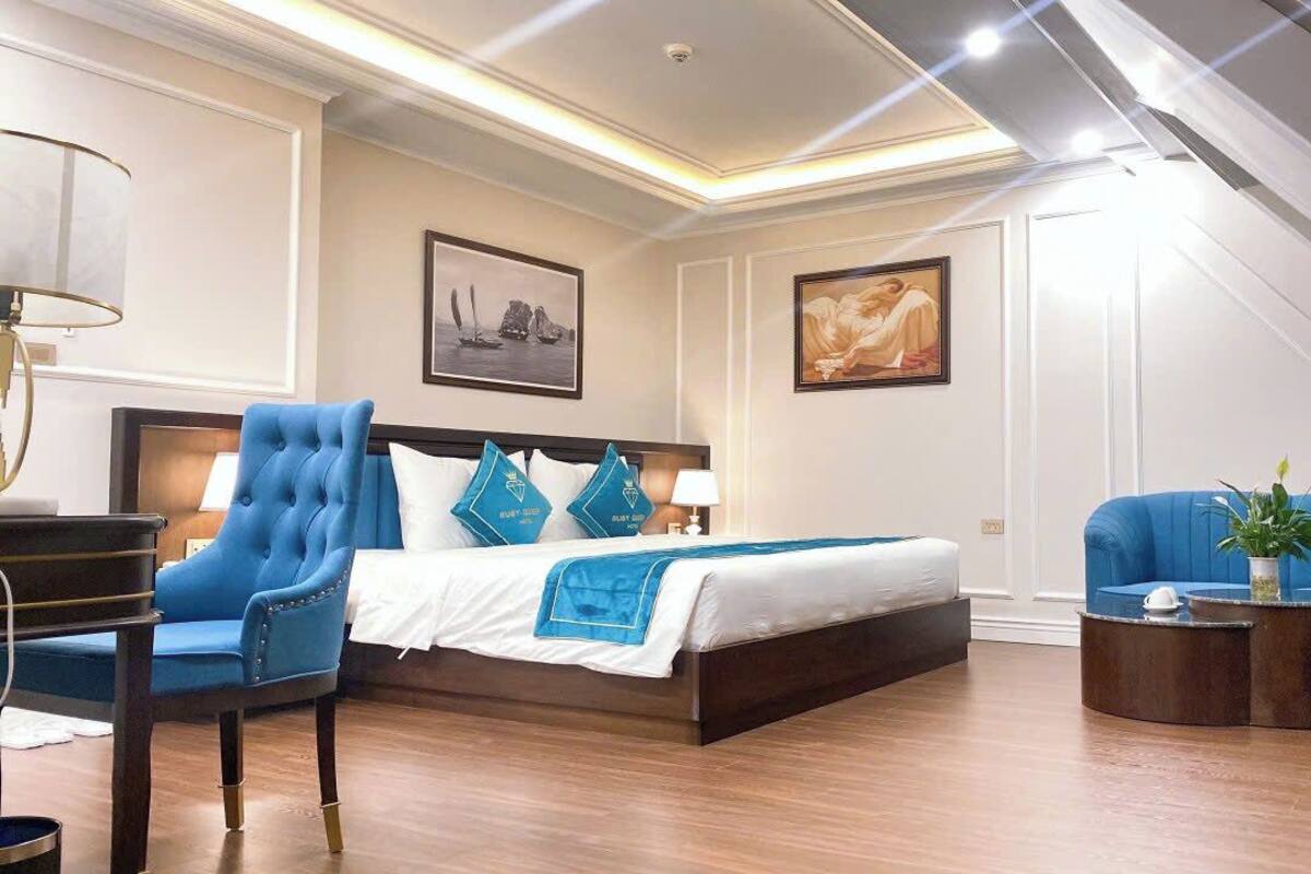 A modern hotel room with a large bed, a blue armchair, a sofa, and a small coffee table.