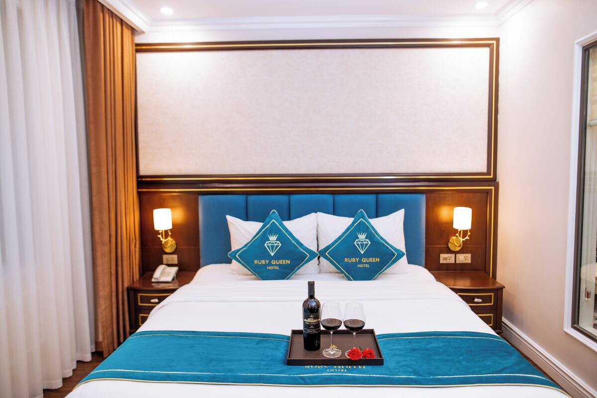 A hotel room with a large bed, two pillows with the hotel's logo, bedside lamps, a tray with a bottle and glasses, and a blue bedspread.