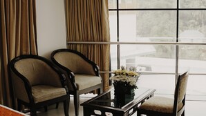 A hotel room with a bed, two armchairs, a small table with a vase of flowers, and large windows with curtains. - Joance Regency (Thodupuzha)
