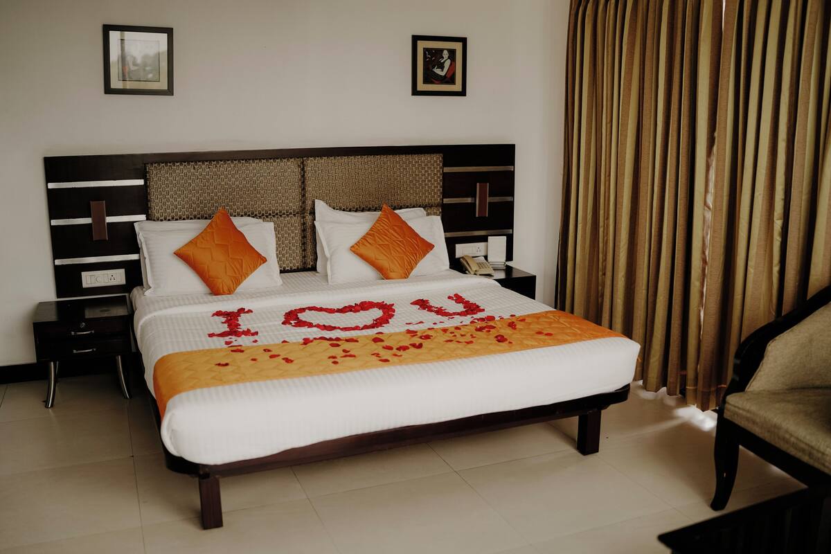 A neatly made bed with a white and orange bedspread, two pillows, and a decorative heart made of rose petals spelling "YOU" on the bed.