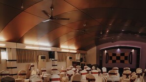 A conference room with rows of chairs covered in white tablecloths and red sashes, a ceiling fan, and a stage with a checkered backdrop. - Joance Regency (Thodupuzha)