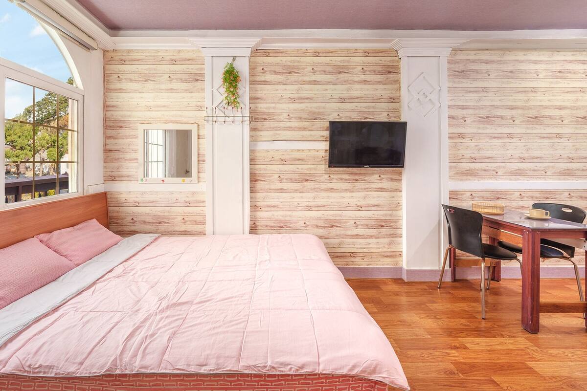 Un dormitorio con cama, televisor, escritorio con silla y ventana con vista a árboles.