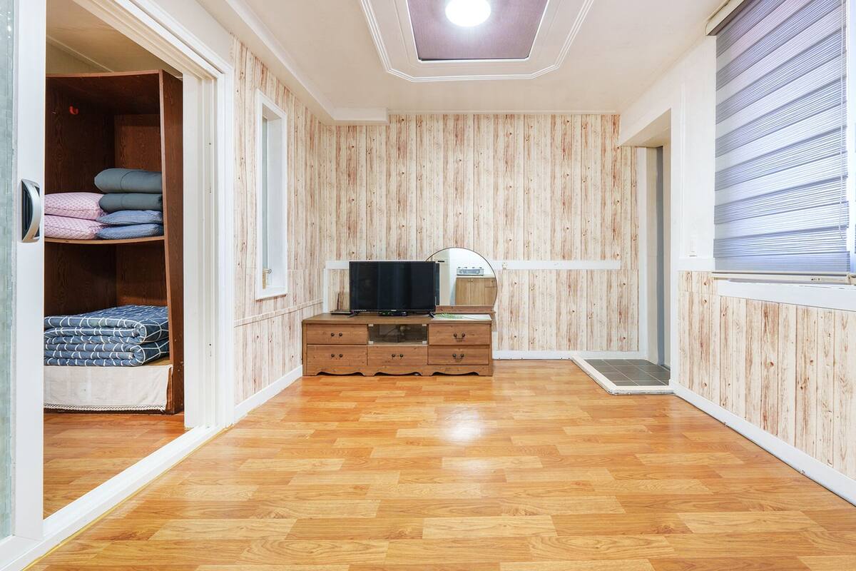 Una habitación con paredes de madera, suelo de madera, un televisor sobre un mueble de madera y una ventana con persianas.