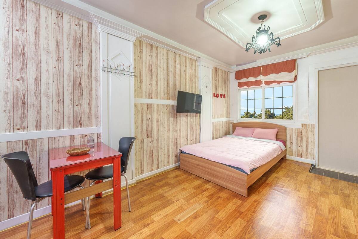 Un dormitorio con piso de madera, una cama con cubrecama rosa, un escritorio con silla, un televisor en la pared y una ventana con vista a árboles.