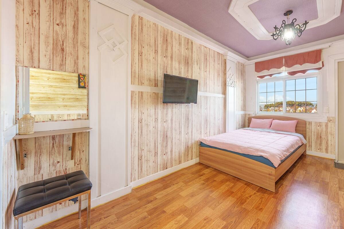 Un dormitorio con pared de madera, una cama con colcha rosa, un televisor montado en la pared y una ventana con vista a casas.