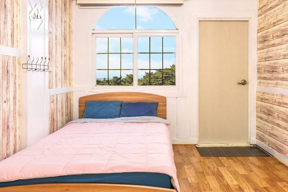 Un dormitorio con cabecera de madera, cama con ropa de cama rosa y azul, ventana con vista a árboles y una puerta.