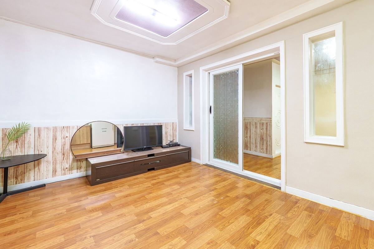 Una habitación con piso de madera, un televisor sobre un mueble de madera, una pequeña mesa con una planta y una puerta corredera de vidrio que da a otra habitación.