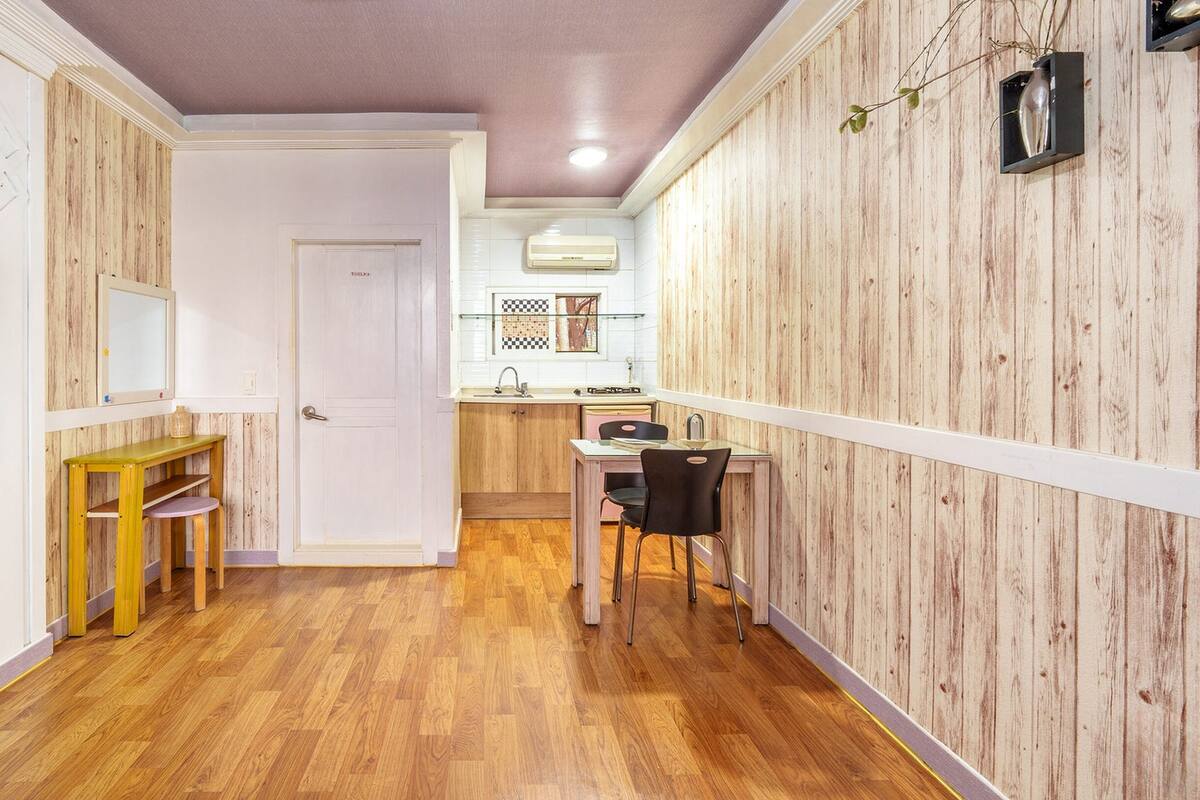 Un pasillo estrecho con paneles de madera, una pequeña mesa de comedor con sillas, un televisor montado en la pared y una puerta que da al baño.