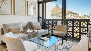 Ein modernes Wohnzimmer mit einer Couch, Sesseln, einem Glastisch und einem Balkon mit Blick auf einen Berg und Gebäude.