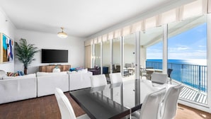 A modern living room with a large TV, a sofa, a dining table, and a balcony with a view of the ocean.