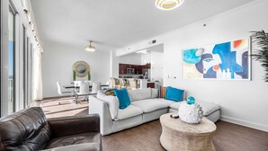 A modern living room with a sectional sofa, a leather armchair, and a round coffee table. There is a dining area with a white table and chairs, and a kitchen in the background.