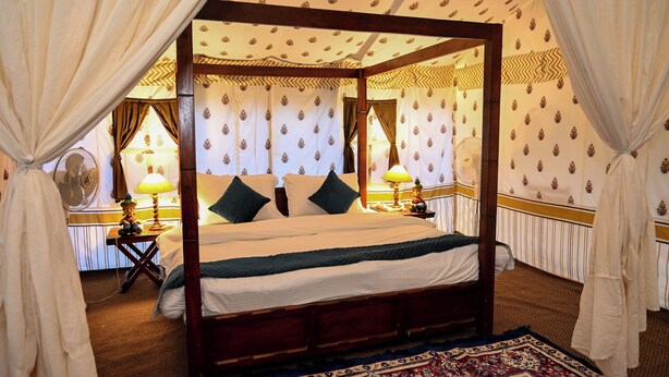Uma cama de dossel com estrutura de madeira, roupa de cama branca e almofadas verdes. Há duas mesas de cabeceira com abajures, um tapete estampado e cortinas.