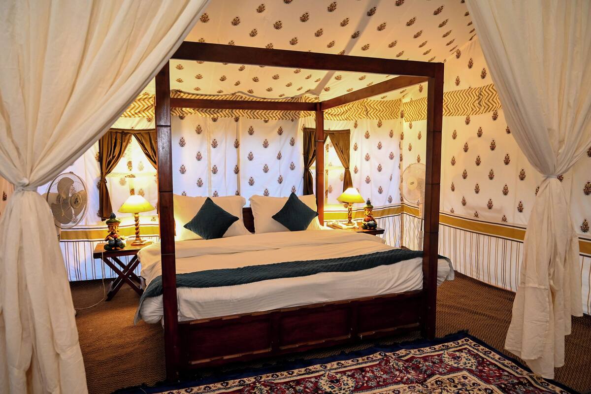 A four-poster bed with a wooden frame, white bedding, and green pillows. There are two bedside tables with lamps, a patterned carpet, and curtains.