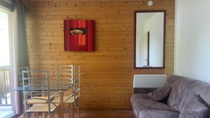 A room with a wooden paneled wall, a glass table with chairs, a brown leather sofa, and a red artwork above the table. - Hameau 3 103 - SPA & Piscine Appartement 6 Pers (Orelle)