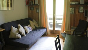 Un salón acogedor con un sofá de color oscuro, suelo de madera, una mesa de comedor con sillas y un ventanal corredero que da a un área exterior.