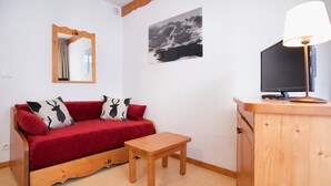 Una habitación con un sofá rojo, una mesa de madera, un televisor sobre un mueble de madera, un espejo y una pintura de un paisaje montañoso.