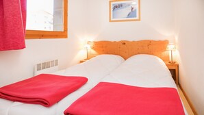 Una cama doble con dos mantas rojas, un cabecero de madera, dos lámparas de noche, un cuadro en la pared y una ventana con vistas a edificios.