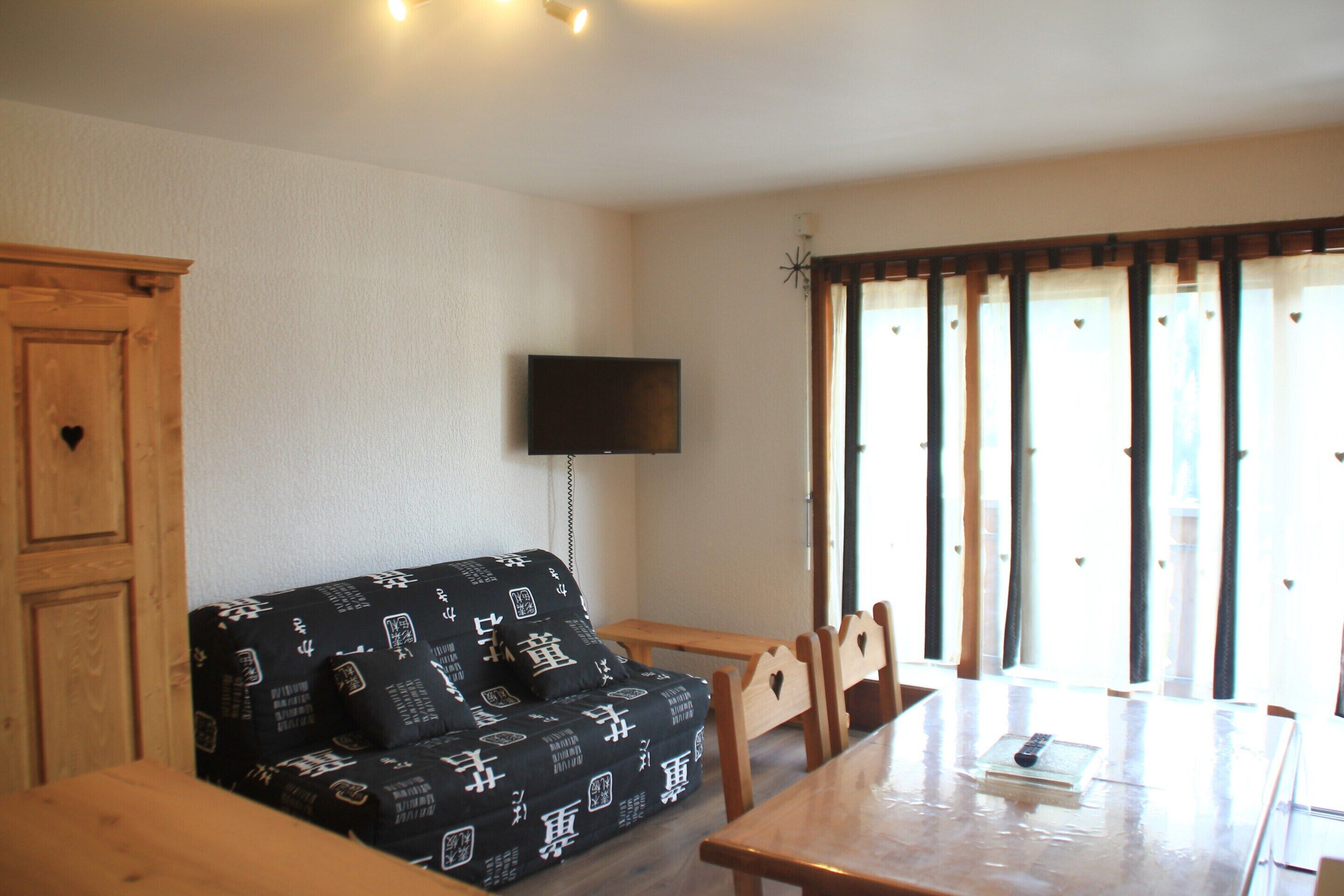 A room with a wooden dining table, a black sofa with patterned cushions, a television mounted on the wall, and a window with curtains.