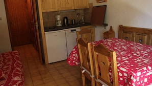 A compact kitchen with wooden cabinets, a sink, and a coffee maker. There is a dining area with a table covered in a red tablecloth and wooden chairs. - Voinettes Vnb2 Telecabine & Mountain 4 Pers (Chatel)