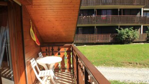 Un balcon en bois, agrémenté d’une table et de chaises, qui offre une vue sur un espace verdoyant et un immeuble à plusieurs étages.