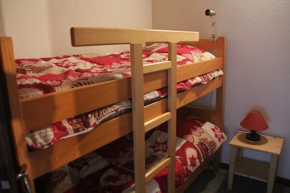 A bunk bed with a red and white patterned bedspread, a small bedside table with a lamp, and a wall-mounted hairdryer.