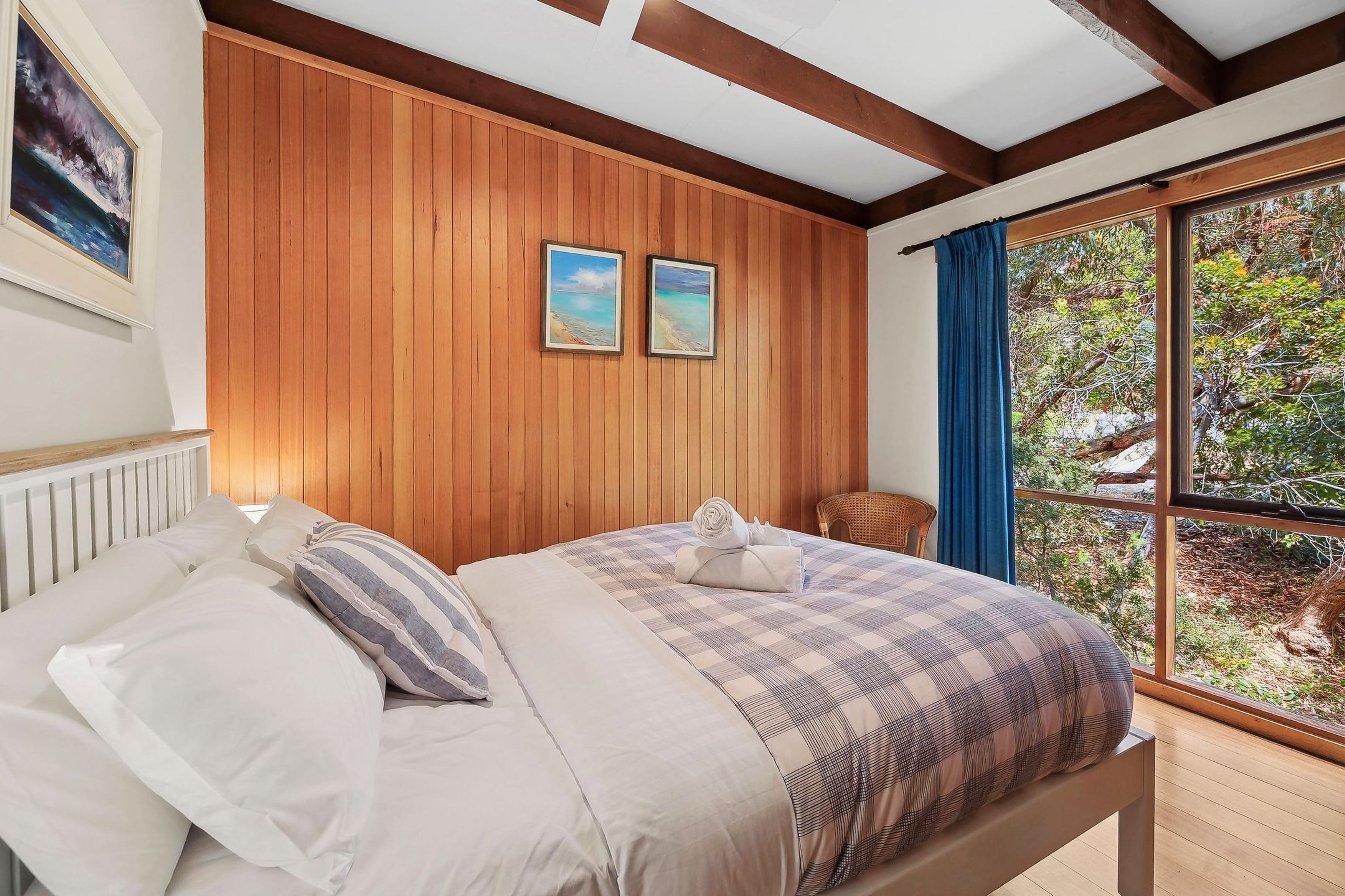 A bedroom with a wooden paneled wall, a bed with a checkered duvet, two framed pictures, a window with a view of greenery, and a wicker chair.