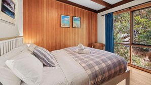 Quarto com parede de painel de madeira, cama com colcha xadrez, duas imagens emolduradas, janela com vista para área verde e uma cadeira de vime.