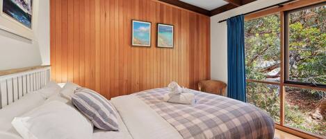 A bedroom with a wooden paneled wall, a bed with a checkered duvet, two framed pictures, a window with a view of greenery, and a wicker chair.