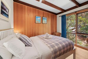 A bedroom with a wooden paneled wall, a bed with a checkered duvet, two framed pictures, a window with a view of greenery, and a wicker chair.