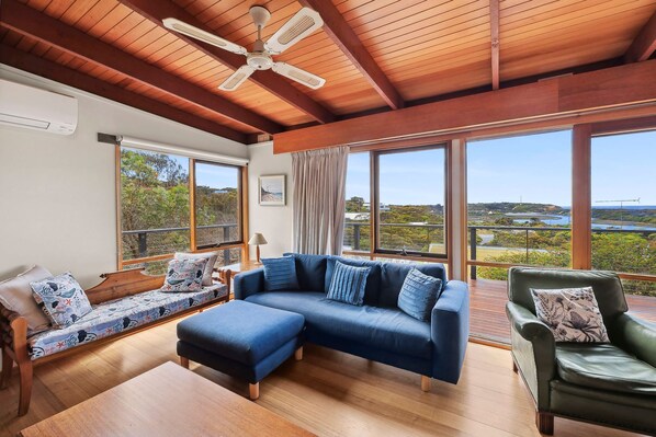 A spacious living area with a wooden ceiling, large windows offering a view of the landscape, a blue sofa, and a green armchair.