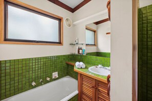 Ein Badezimmer mit einer Badewanne, einem Waschbecken und einem Spiegel. Die Wände sind mit grünen Fliesen versehen und über dem Waschbecken befindet sich ein Fenster.