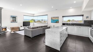 A modern kitchen with a marble island, dark flooring, and large windows offering views of the outdoors.