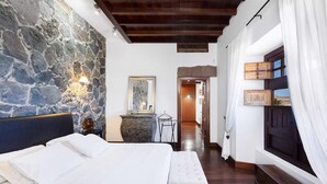 Un dormitorio con pared de piedra, cama, banco, cómoda y ventana con cortinas.