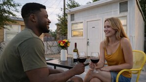 Deux personnes sont assises à une table à l’extérieur, devant des verres à vin, une bouteille de vin et un vase de fleurs.